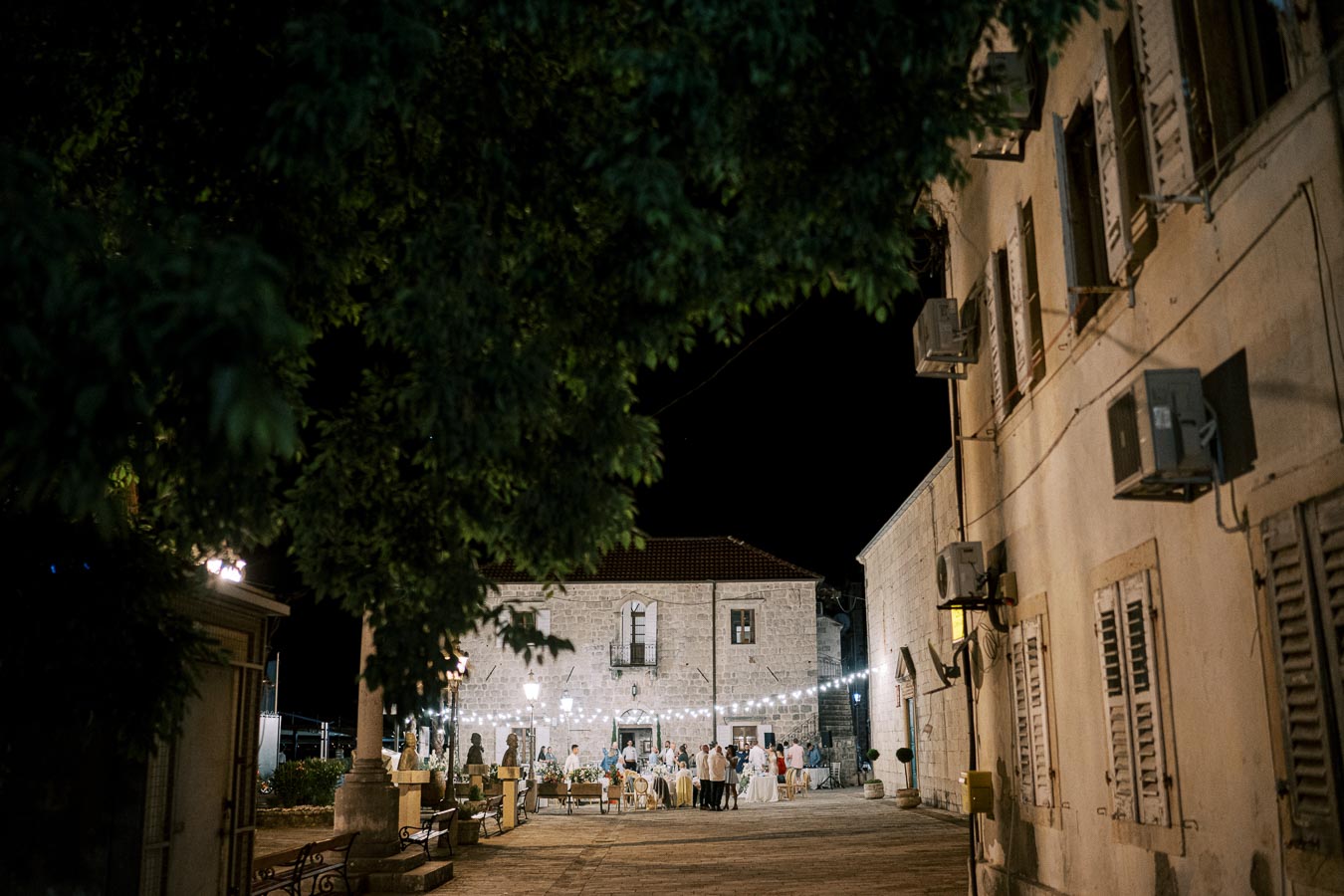 Outdoor night event in a quaint cobblestone courtyard, illuminated by string lights, surrounded by historic stone buildings with wooden shutters and air conditioning units.