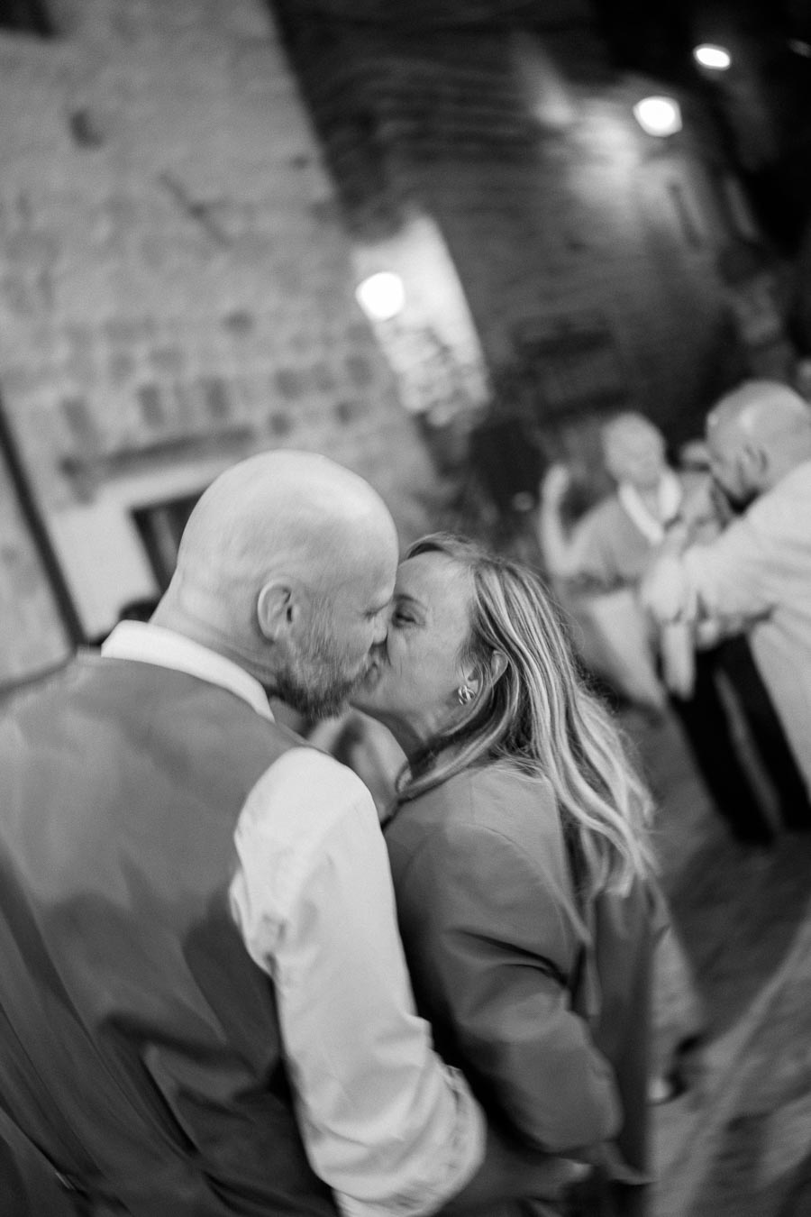 Black and white photo of a couple sharing a kiss on a cobblestone street at night, with blurred lights in the background creating a romantic atmosphere.