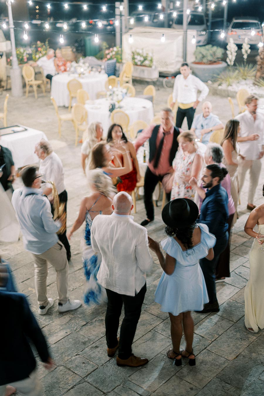 A lively outdoor wedding reception with elegantly dressed guests dancing under string lights, while a musician plays the saxophone amidst a festive atmosphere.