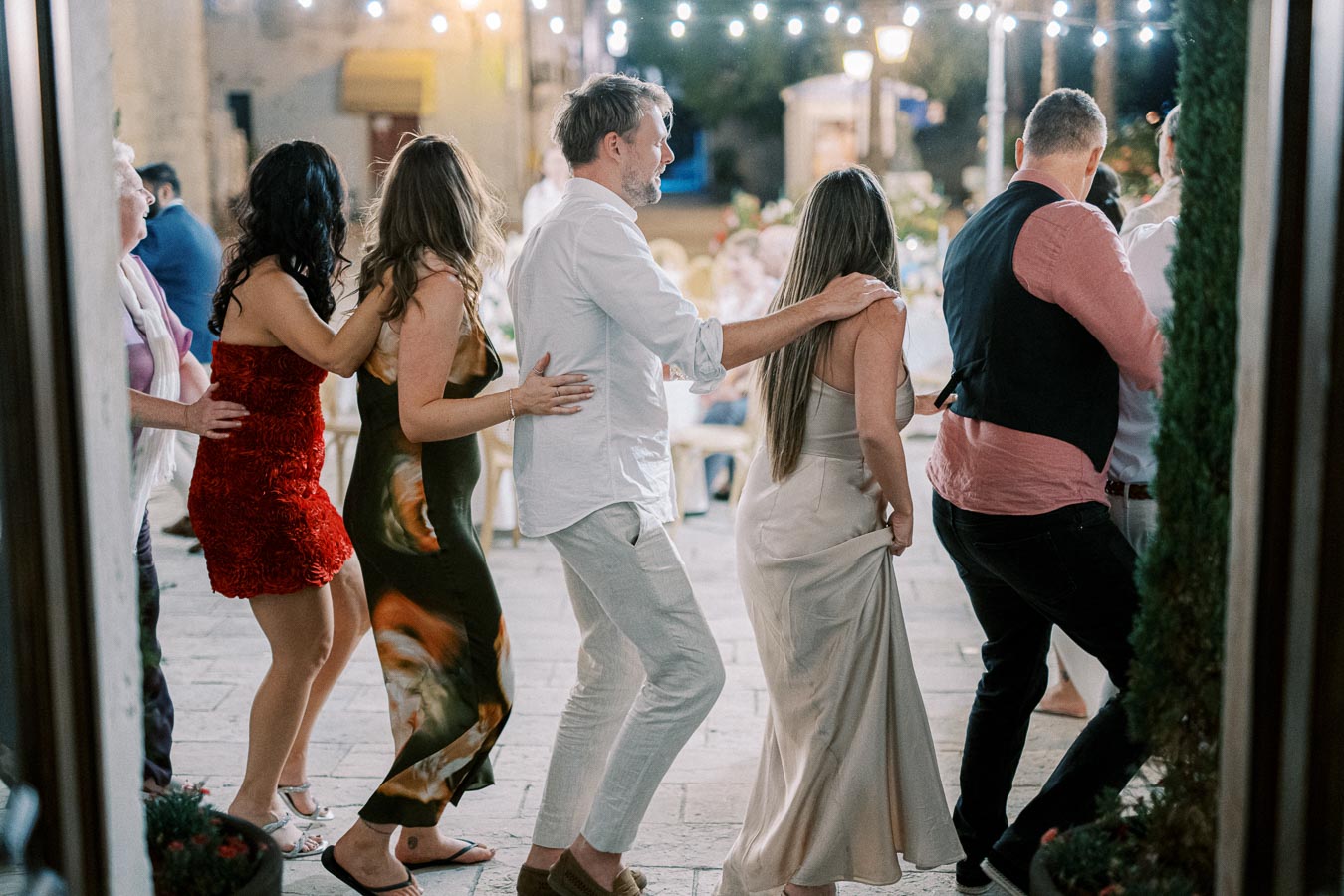 A group of people enjoying a lively conga line at an outdoor evening event, with festive lights illuminating the scene.
