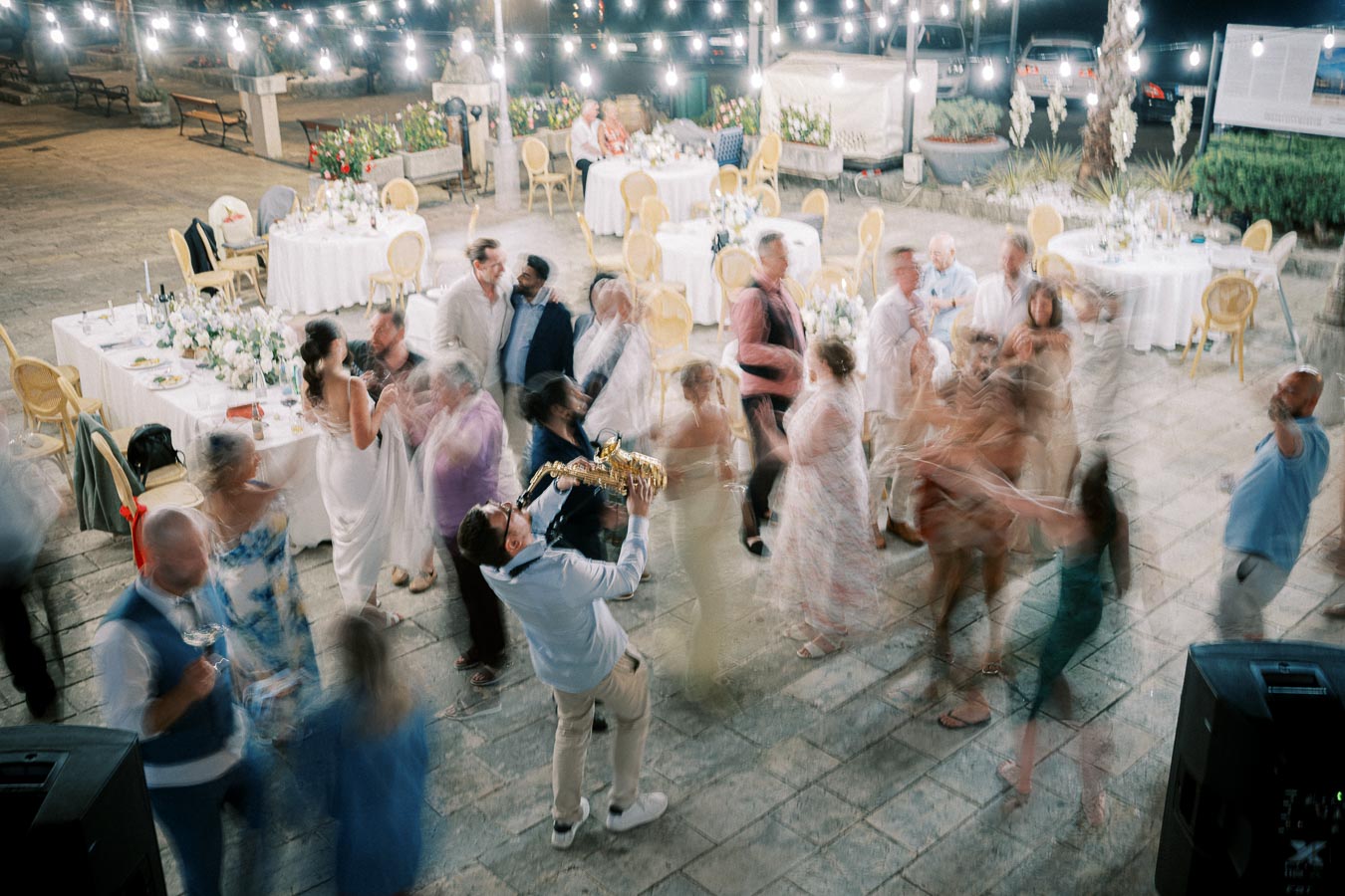 A vibrant wedding reception with guests dancing energetically under string lights. An enthusiastic musician plays the saxophone in the center, surrounded by elegantly set tables adorned with flowers and chairs.