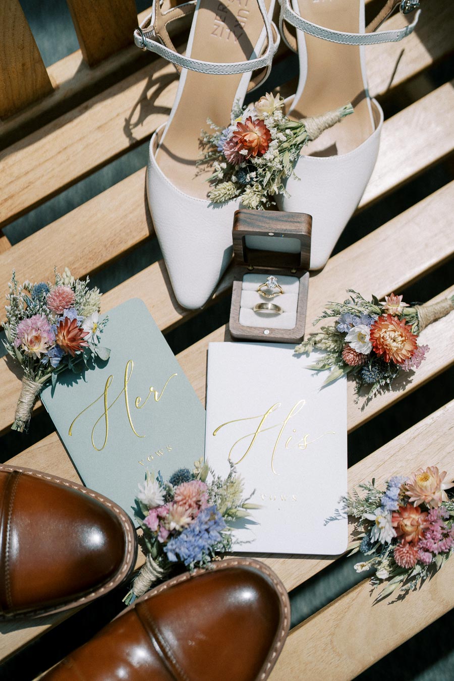 Wedding preparation flat lay featuring bridal shoes, groom's shoes, floral boutonnieres, His and Her vow booklets, and a ring box with wedding rings on a wooden background. Ideal for wedding inspiration and planning themes.