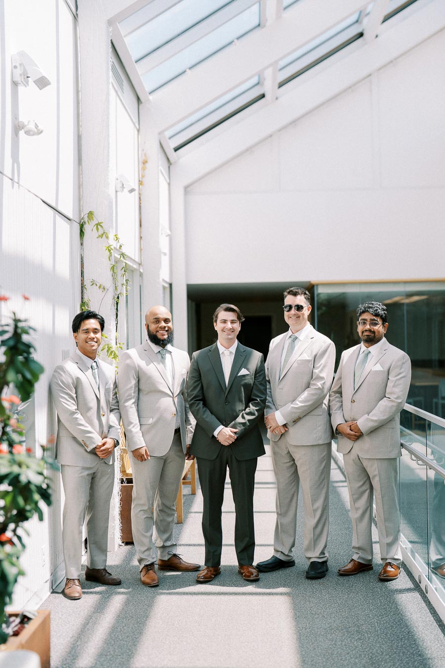 A group of five men in suits standing in a modern, sunlit corridor with glass walls and plants nearby. The men are smiling, exuding a sense of professionalism and camaraderie, ideal for a business or formal event setting.