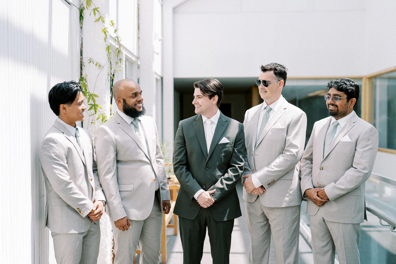 A group of five men in suits, including one in a dark suit and the rest in light suits, standing indoors and smiling, with natural light streaming from above creating a bright and airy atmosphere.