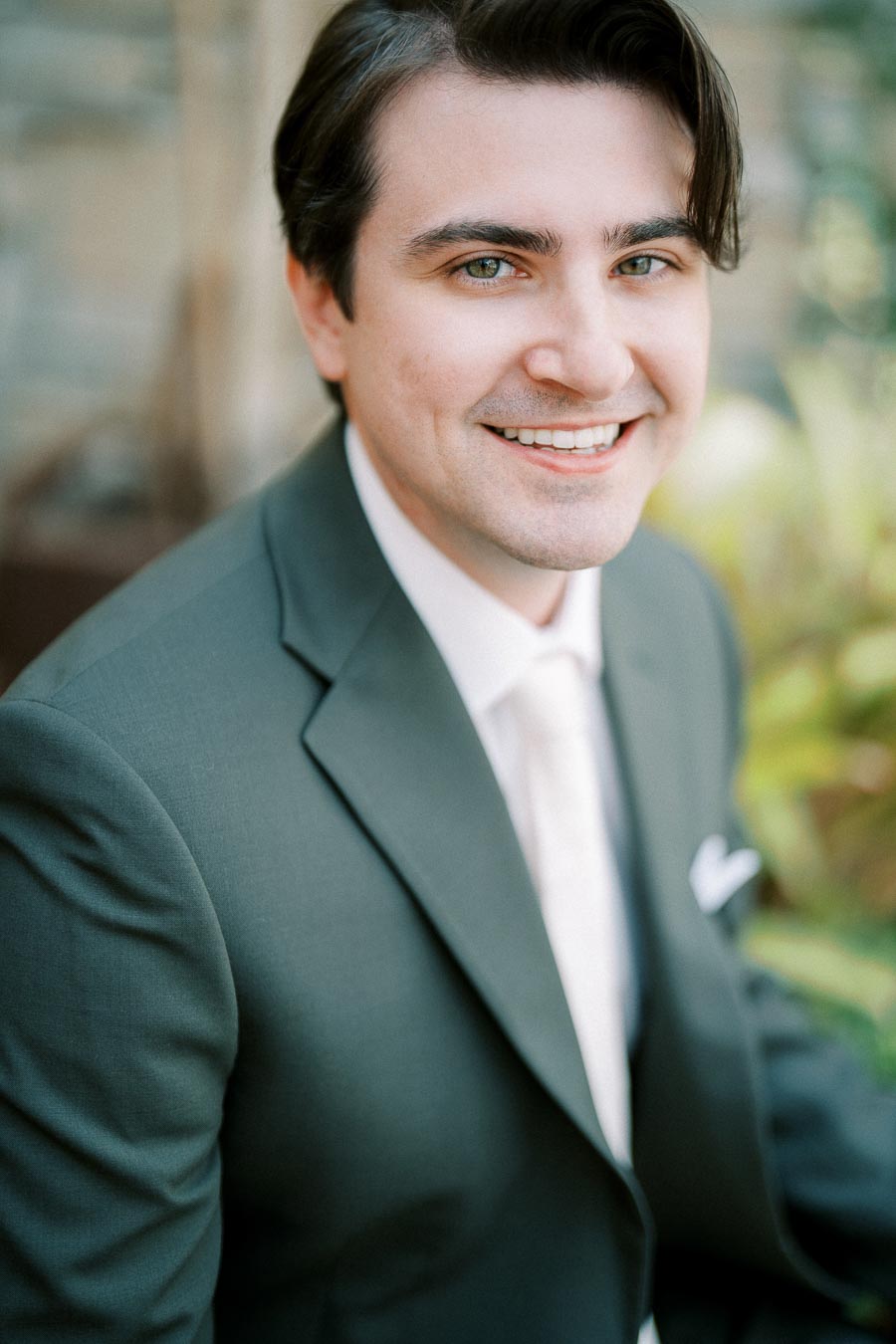 Sure! Here's a suitable alt text:Smiling man in a green suit with a white shirt, posing outdoors with a blurred leafy background.