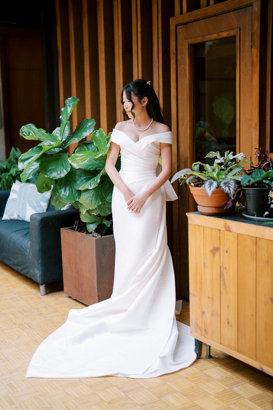 A bride in an elegant off-the-shoulder white wedding gown stands in a stylish indoor setting with lush green plants and wooden decor.
