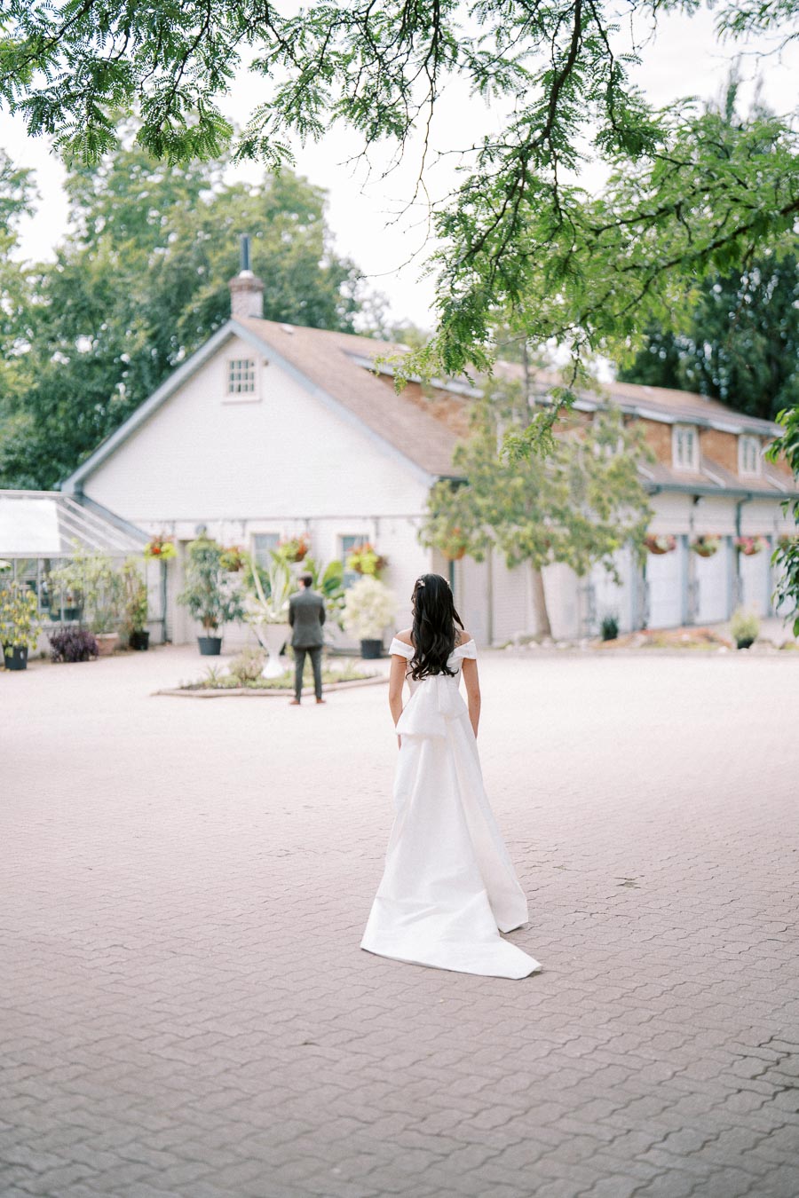 A bride in a white dress approaches a groom standing in a courtyard outside a rustic building, surrounded by greenery and hanging plants.