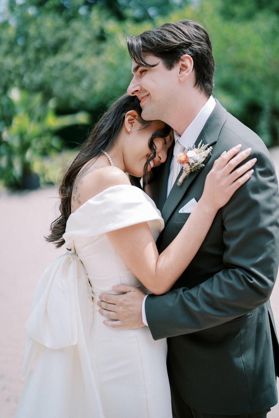 Bride and groom embracing in a lush garden, the bride in an elegant off-shoulder wedding dress and the groom in a dark suit, both smiling peacefully.