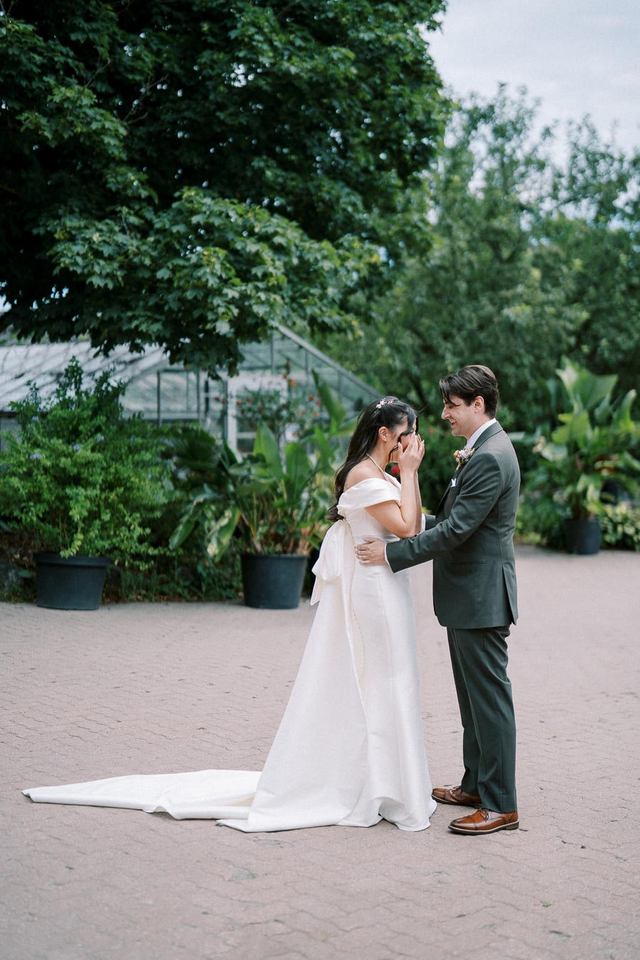 A joyful couple shares an emotional moment during their wedding day, standing in a beautiful garden setting with lush greenery and a greenhouse in the background. The bride, in an elegant white gown, covers her face with her hands, while the groom, in a formal suit, lovingly embraces her.