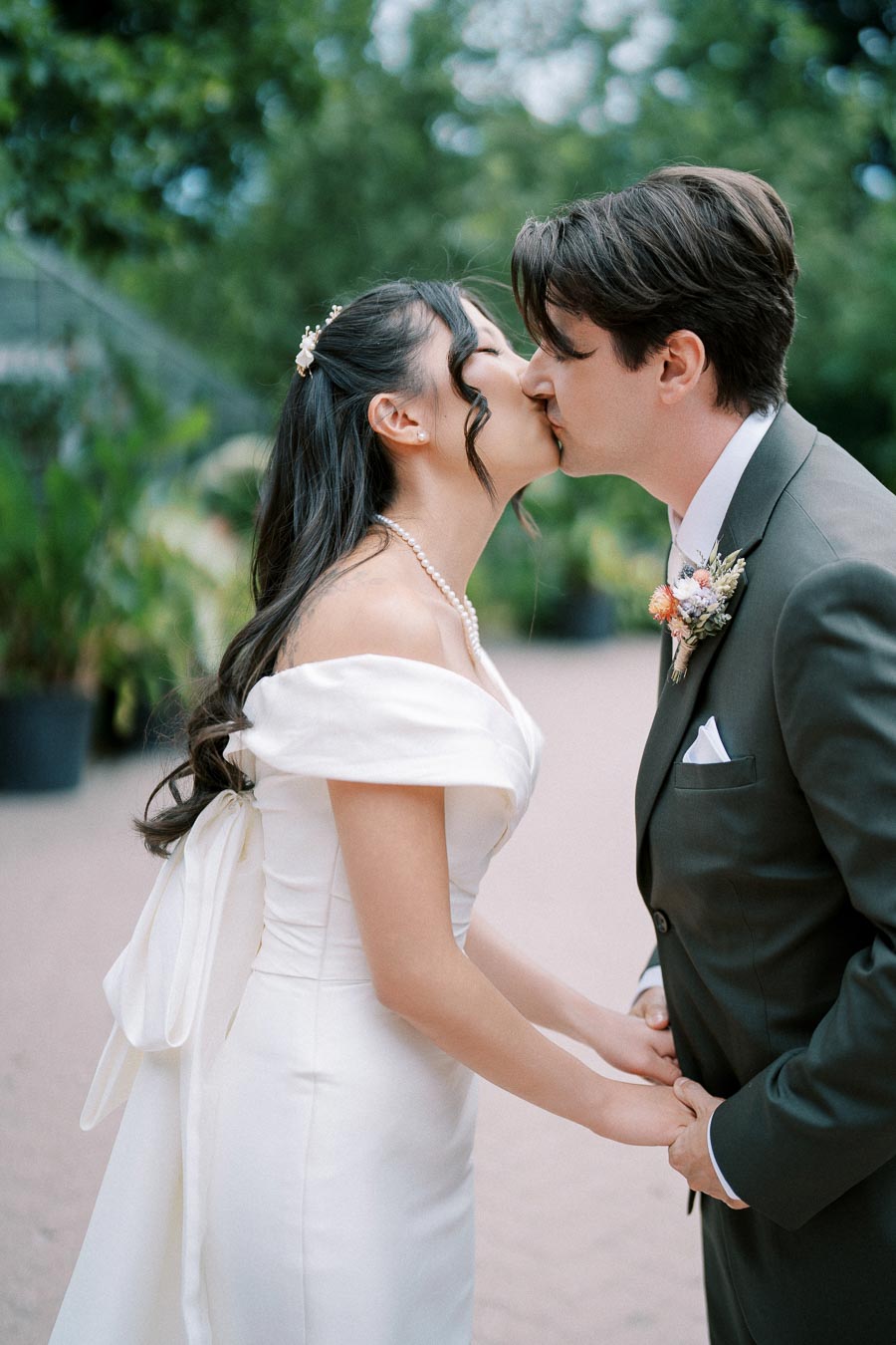 A bride and groom in elegant wedding attire share a romantic kiss outdoors, surrounded by lush greenery.