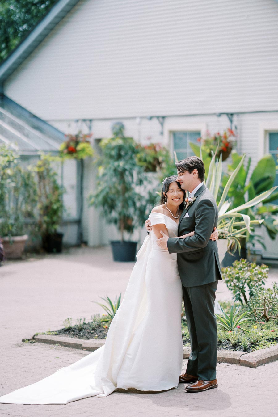 A newlywed couple embracing and smiling in a garden setting in front of a white building, with greenery and flowers in the background. The bride is wearing a white wedding gown with a long train, and the groom is in a dark suit.