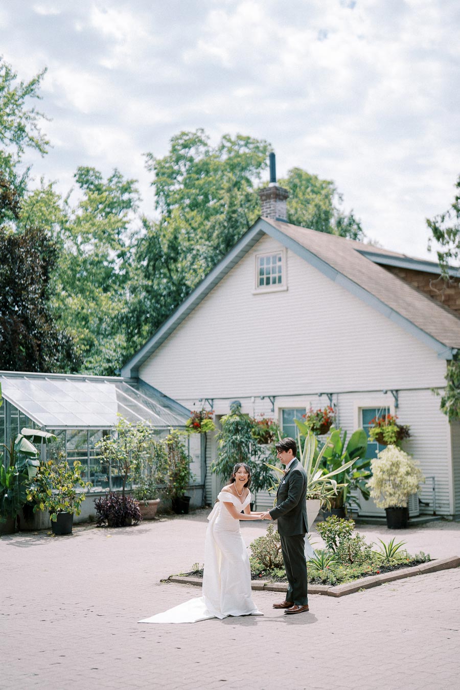 Bride and groom sharing a joyful moment outside a charming greenhouse with lush greenery and a rustic farmhouse backdrop.