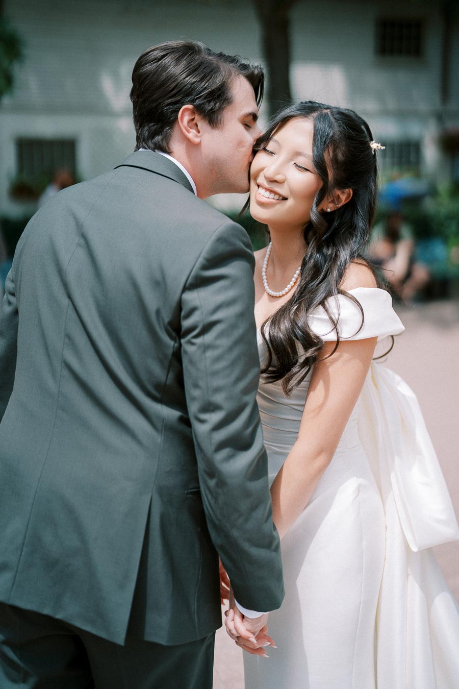 A groom in a formal suit kisses the bride's cheek as they hold hands, with the bride in a white wedding dress smiling under outdoor sunlight.