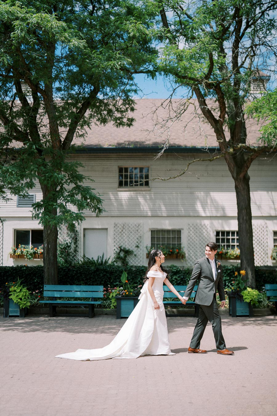 A bride and groom holding hands while walking outdoors in front of a charming white house, surrounded by lush green trees and colorful flowers under a clear blue sky.