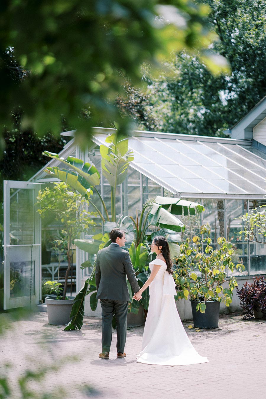 A bride and groom holding hands in front of a lush greenhouse, surrounded by greenery and potted plants, creating a serene and romantic wedding atmosphere.