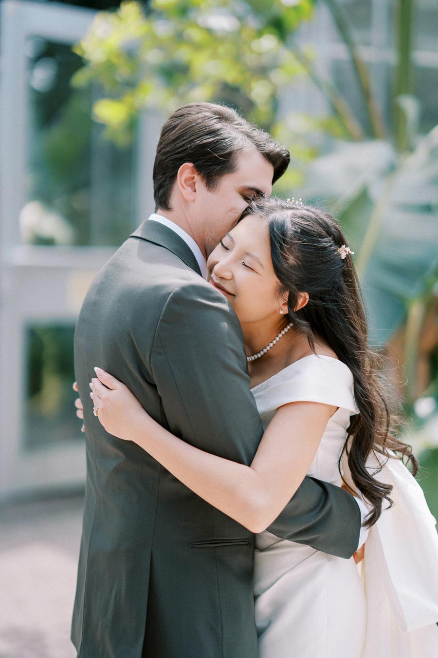 A bride and groom embrace lovingly in an outdoor wedding setting, surrounded by lush greenery and natural light. The bride wears an elegant white gown, and the groom is in a dark suit, creating a romantic atmosphere.