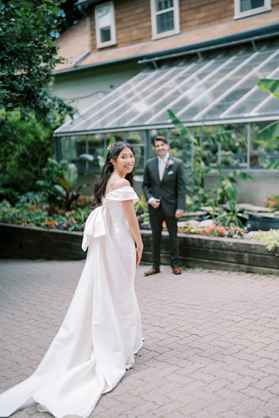 A bride in an elegant white gown smiles as she looks at her groom standing behind her. The scene is set in a lush garden with a glass greenhouse in the background, creating a romantic and picturesque wedding moment.