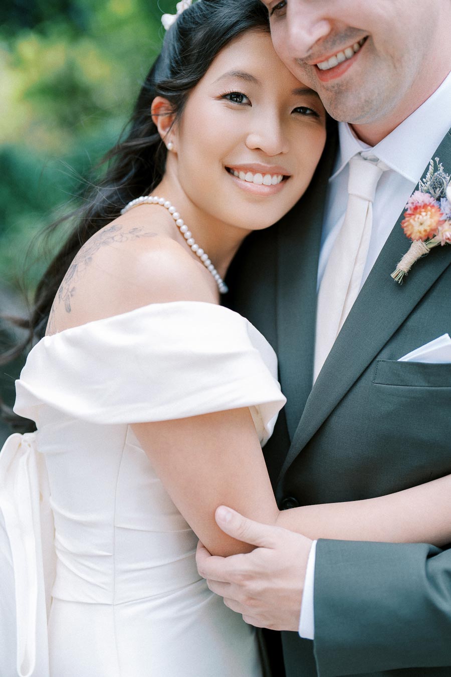A smiling bride in an off-the-shoulder wedding dress with pearl necklace embraces her groom in a gray suit, showcasing a tender moment in a lush outdoor setting.