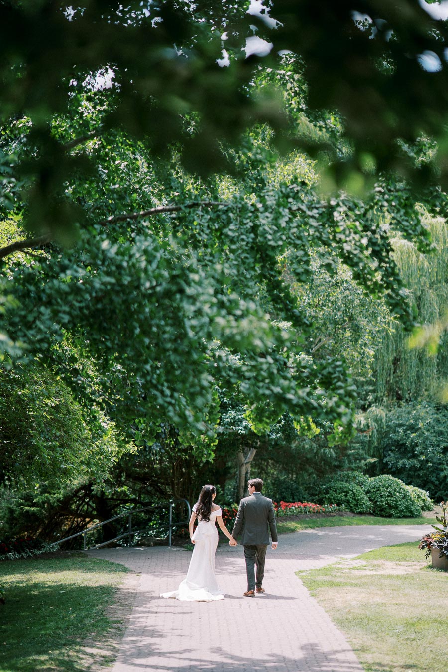A bride and groom walking hand in hand down a tree-lined path in a lush, green garden on a sunny day.