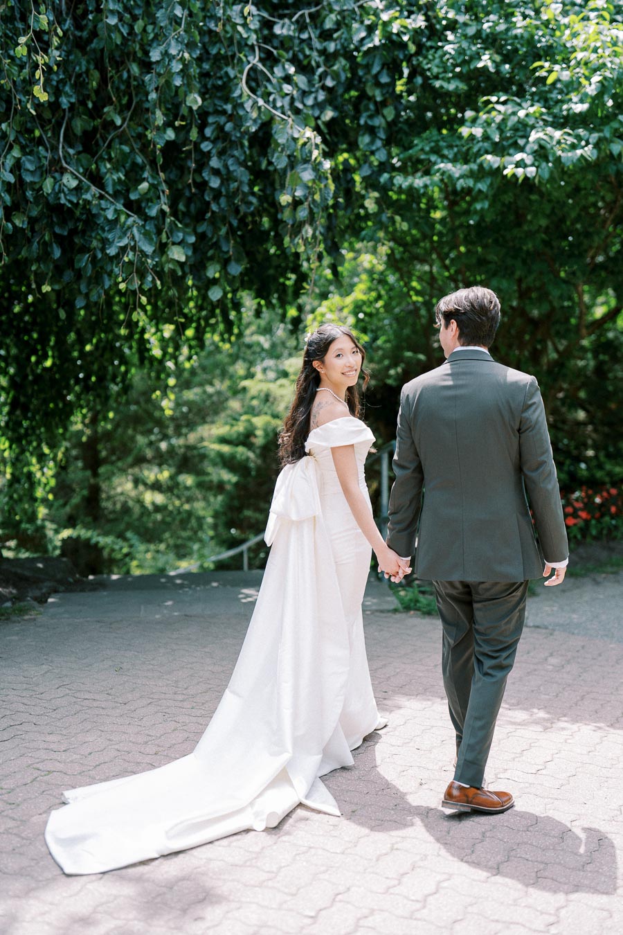 A bride in a white dress and a groom in a suit hold hands while walking through a lush garden, surrounded by vibrant greenery.