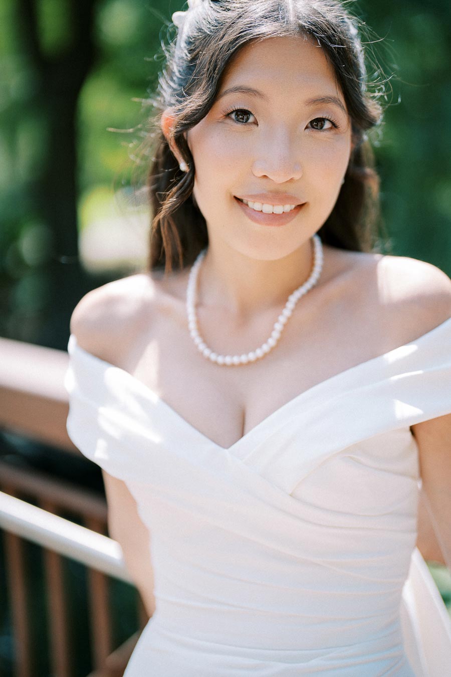 A bride smiling in a white off-shoulder wedding dress with a pearl necklace, surrounded by a green outdoor setting.