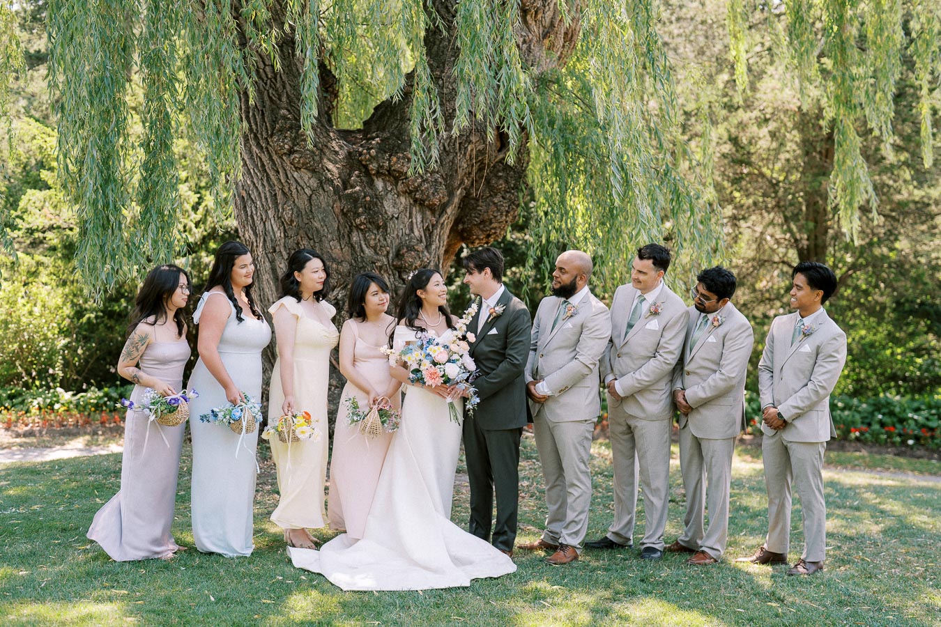 A wedding party poses outdoors under a large tree. The bride in a white gown and groom in a dark suit stand in the center, surrounded by bridesmaids in pastel dresses and groomsmen in light suits. They are all smiling, holding bouquets, with lush greenery in the background.