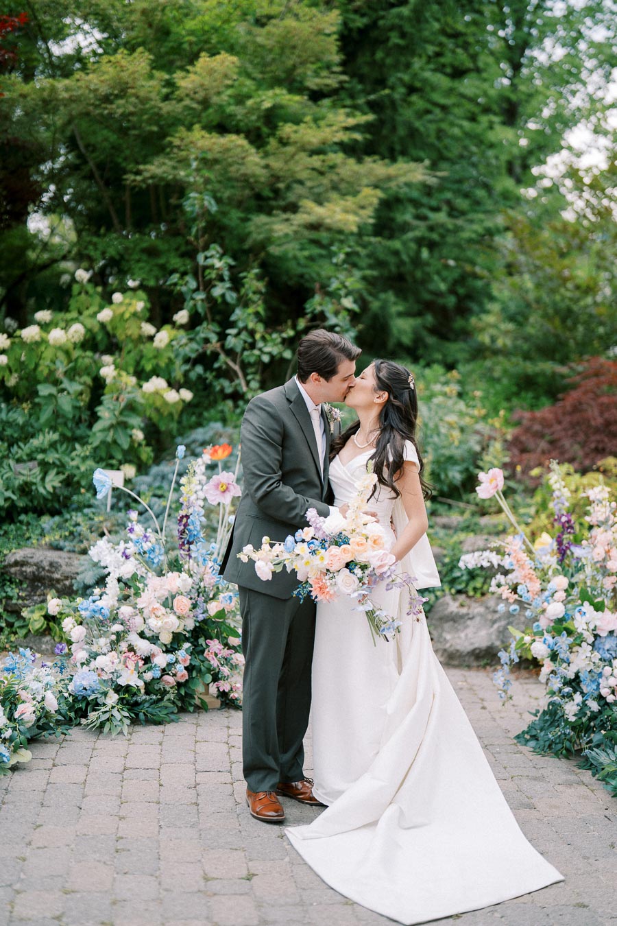 A bride and groom kiss in a lush garden setting, surrounded by colorful floral arrangements. The bride is in a white gown holding a bouquet of soft pastel flowers, while the groom wears a dark suit, providing a romantic and picturesque wedding scene.