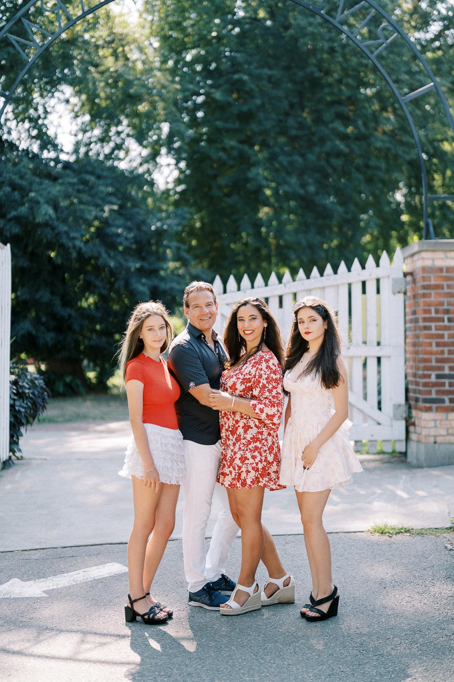 A family of four posing together outdoors near a white picket fence, with greenery in the background, dressed in summer outfits.