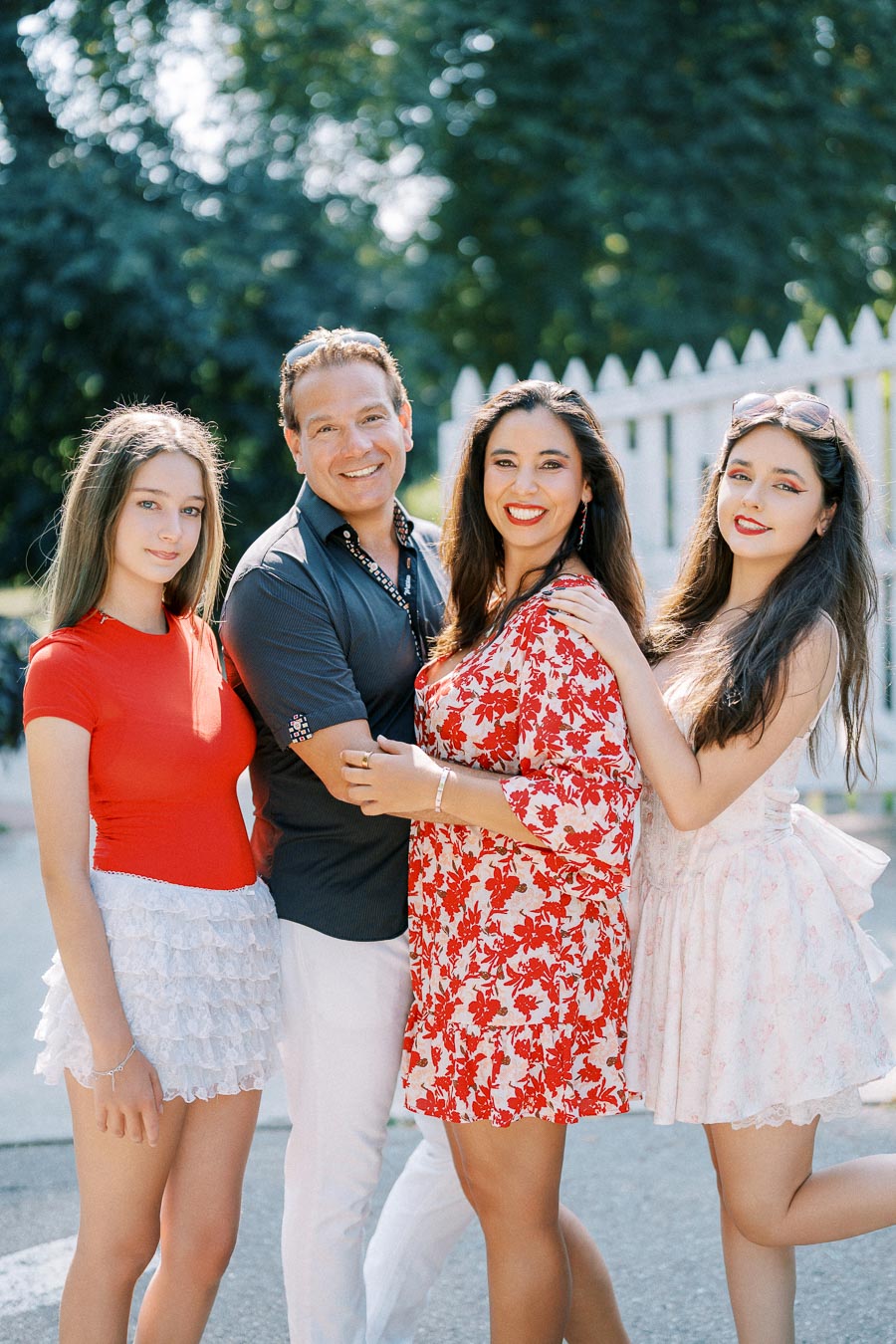 Alt text: A happy family of four posing outdoors, featuring two parents and their teenage daughters. The mother wears a red floral dress, while the father is in a dark shirt and white pants. The daughters are dressed in vibrant outfits, standing together in front of a white picket fence with greenery in the background, suggesting a sunny day.