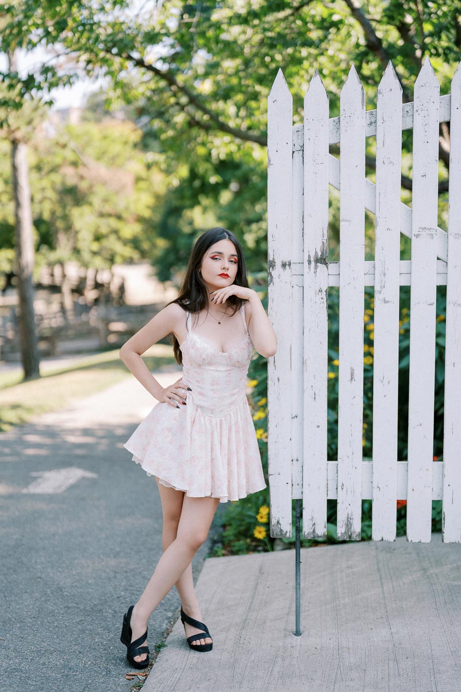 Young woman in a floral dress posing by a white picket fence in a sunny park setting.