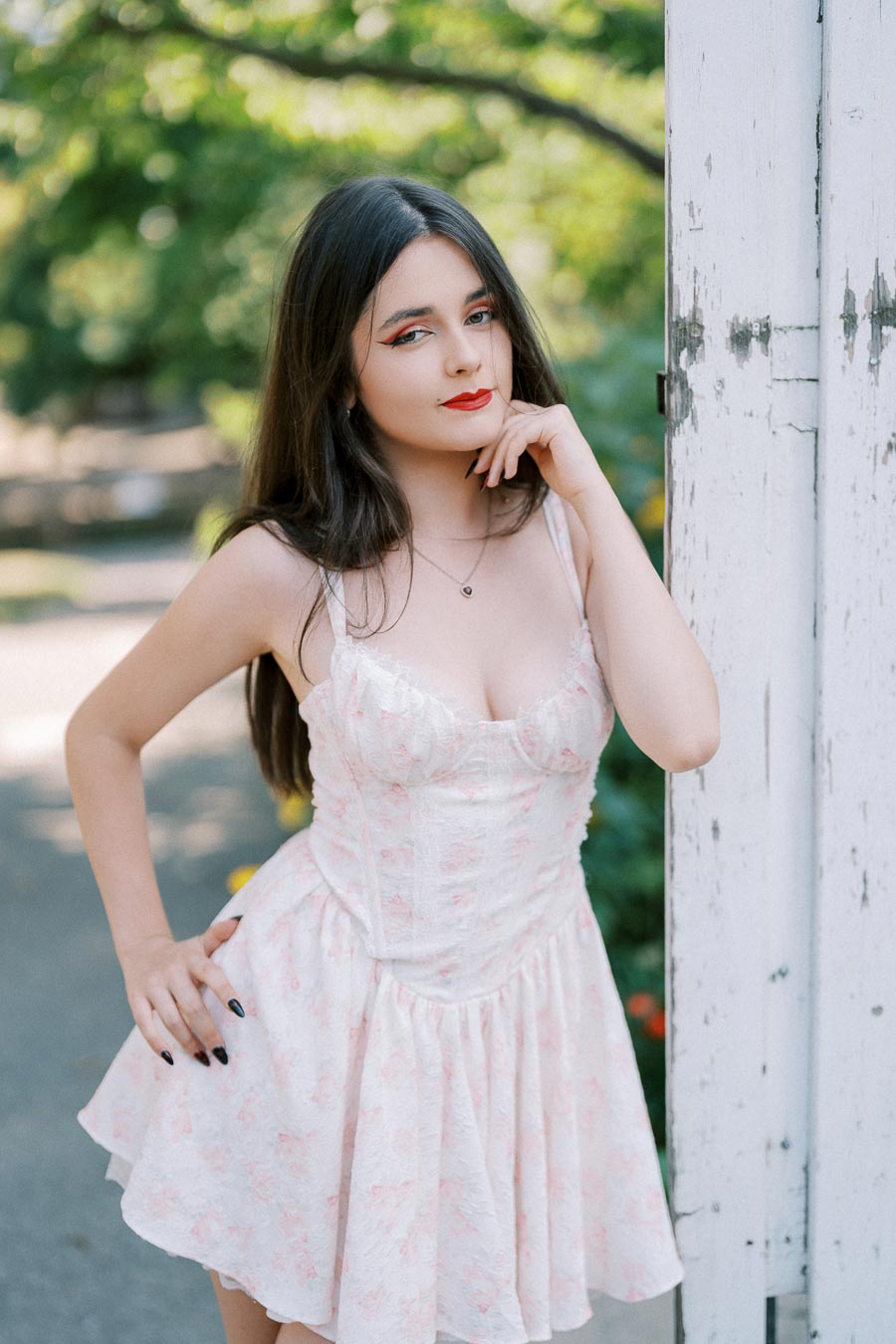 Young woman in a floral dress posing outdoors against a rustic white wooden door, with lush greenery in the background.