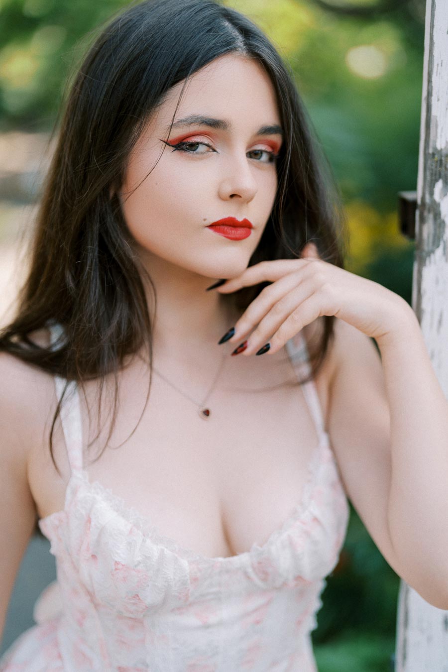 ```plaintext Portrait of a woman with long dark hair, wearing red lipstick and eye makeup, in a light floral dress, posing outdoors with a thoughtful expression. ```