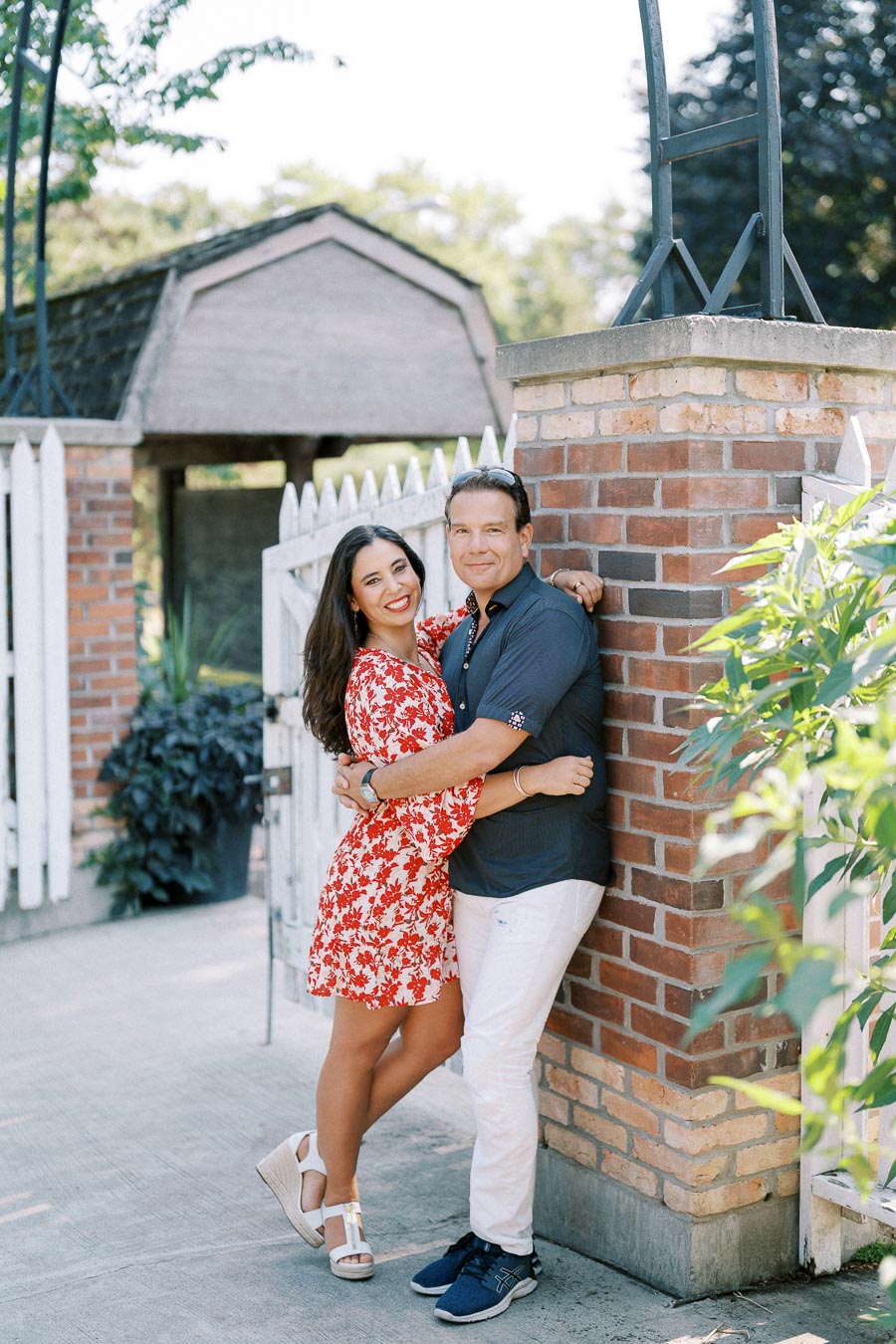 A couple embraces in a garden setting, with a white picket fence and brick wall behind them. The woman wears a red floral dress and white sandals, while the man wears a navy shirt and white pants. They are smiling and look relaxed outdoors.