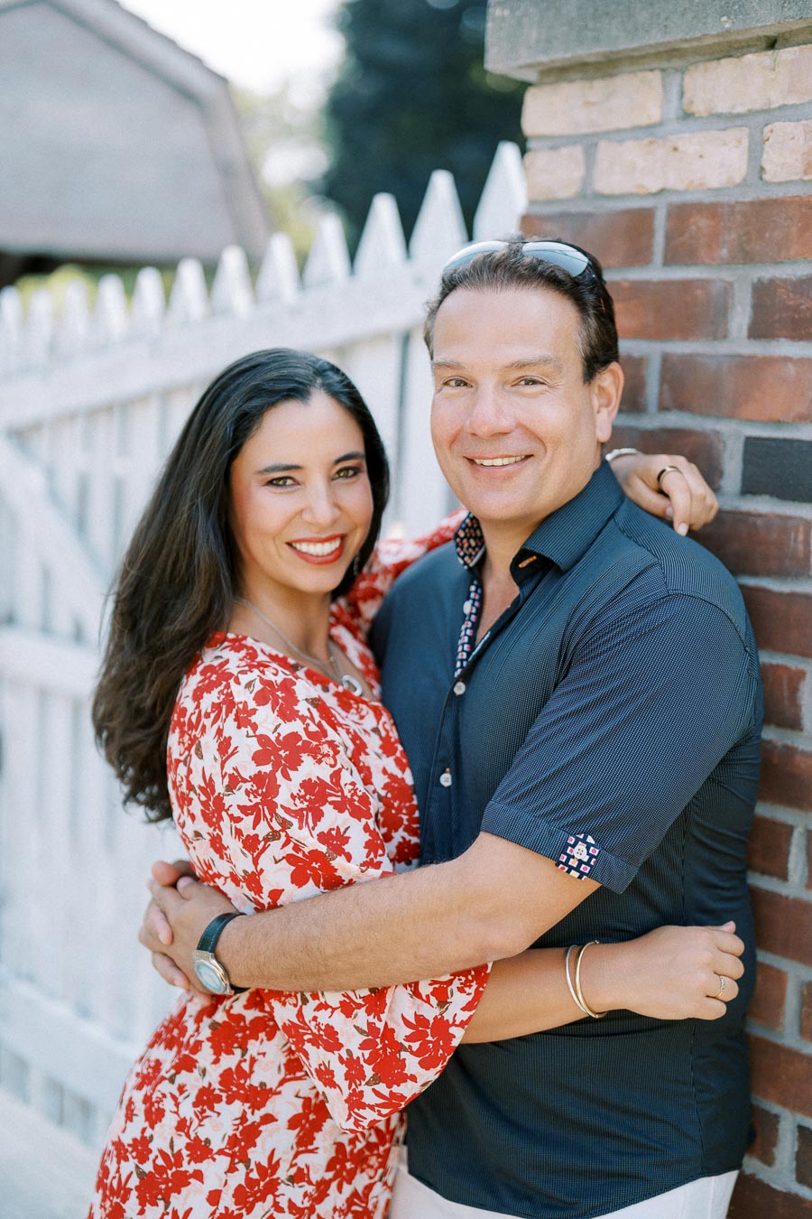 A smiling couple embracing in front of a brick wall and white picket fence on a sunny day. The woman is wearing a red floral dress, and the man is wearing a dark blue shirt.