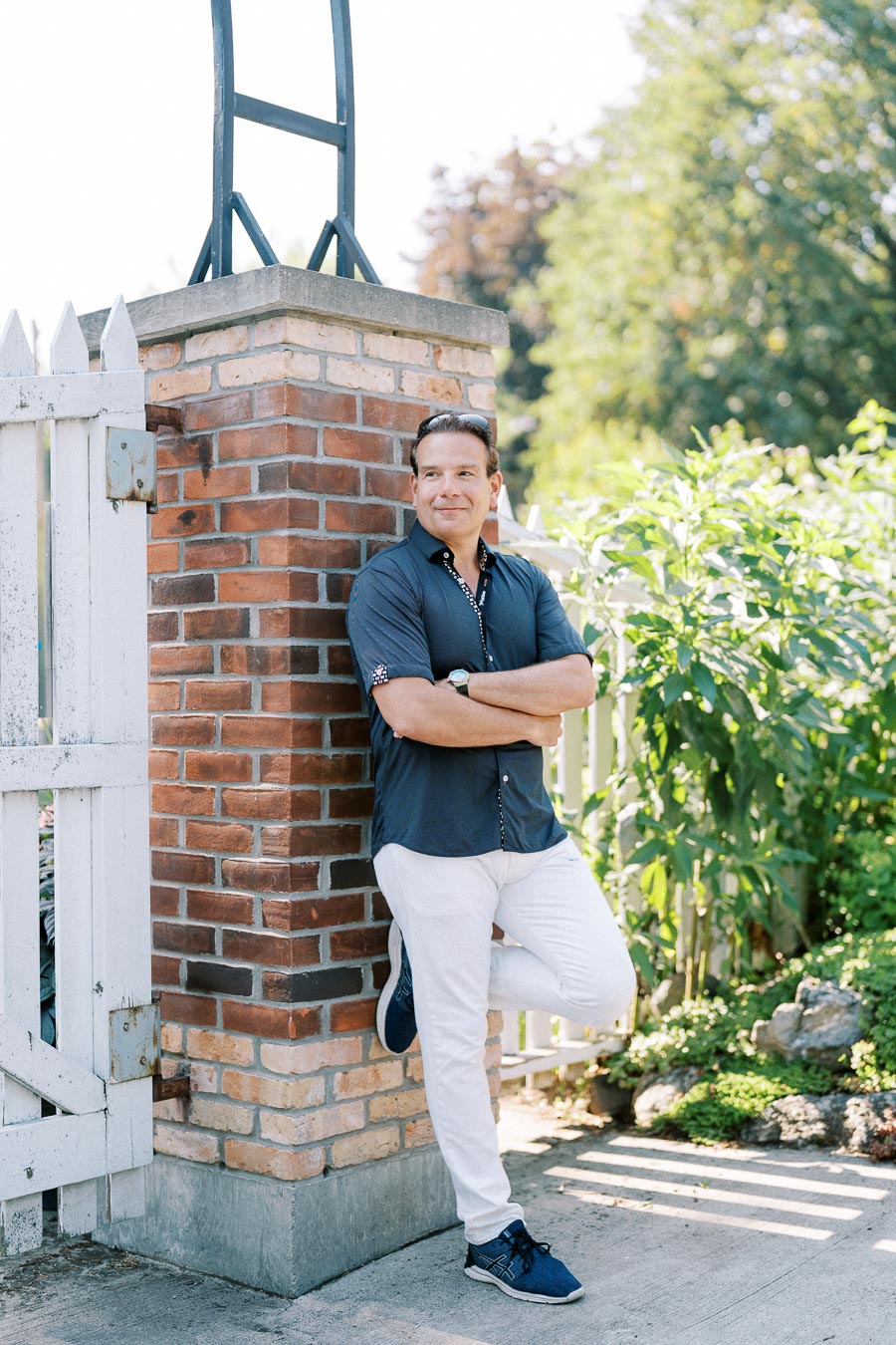 Man in casual attire leaning against a brick pillar, surrounded by greenery and a white fence, in a sunny outdoor setting.
