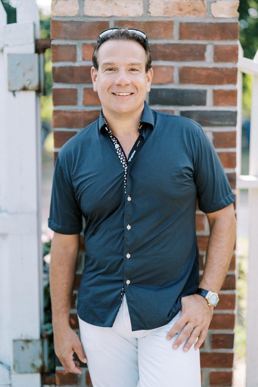 Certainly! Here’s an SEO-friendly alt text for the image:Smiling man in navy blue shirt leaning against a brick wall on a sunny day.