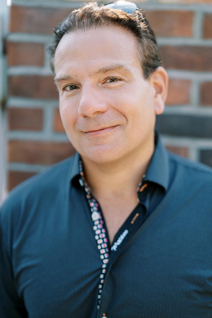 Sure! Here's an SEO-friendly alt text for the image:Smiling man in a blue shirt standing in front of a brick wall.