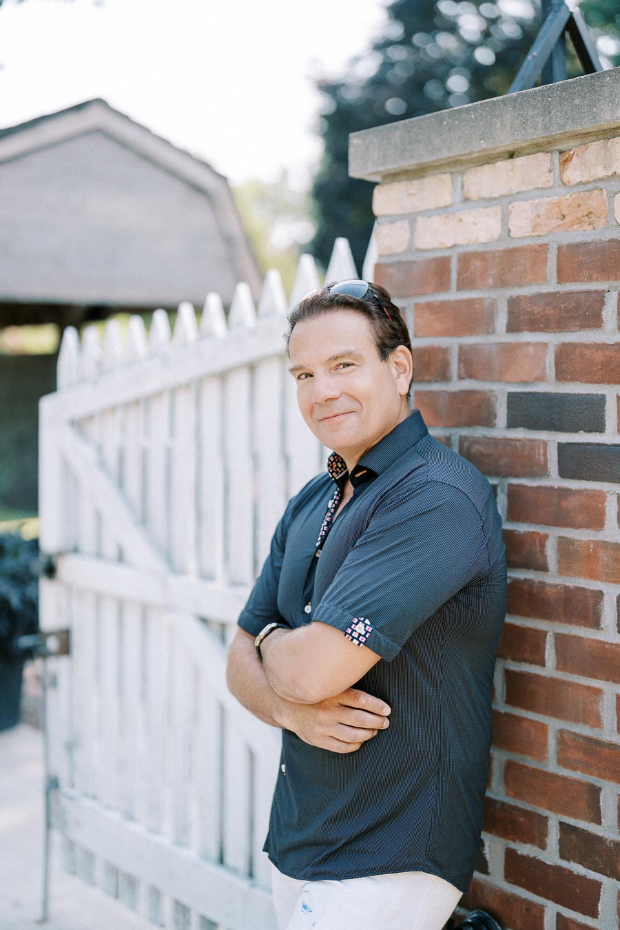 Man in a blue shirt stands confidently against a red brick wall next to a white picket fence, outdoors on a sunny day.