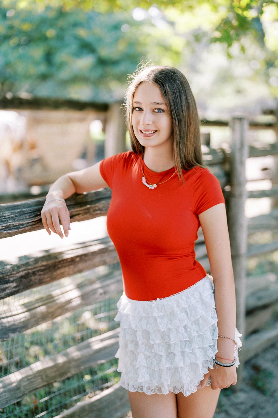 Young woman in a vibrant red top and white lace skirt standing by a rustic wooden fence, surrounded by lush greenery and sunlight.