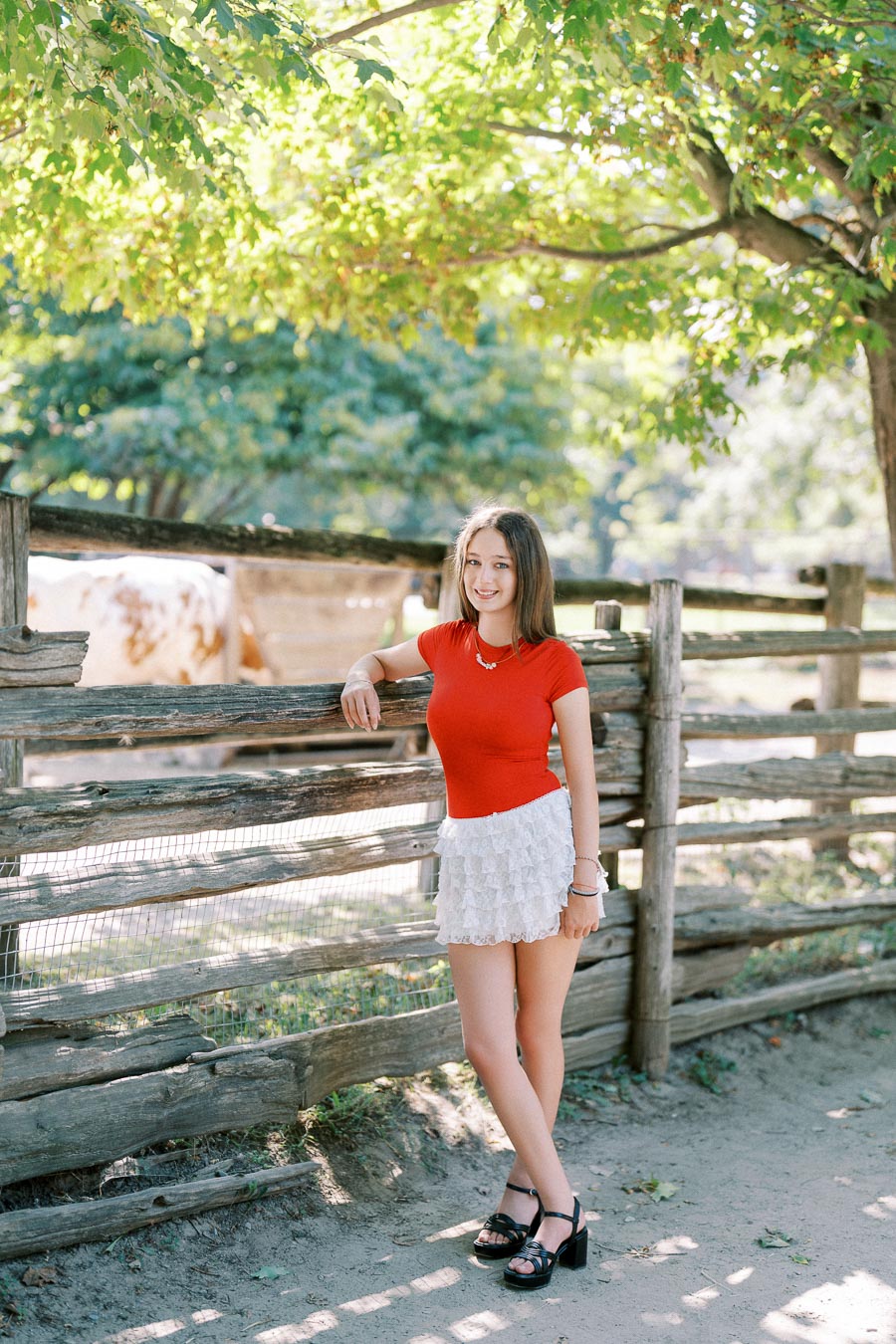 Young woman in a red top and white skirt smiling by a rustic wooden fence on a sunny day, surrounded by leafy green trees.