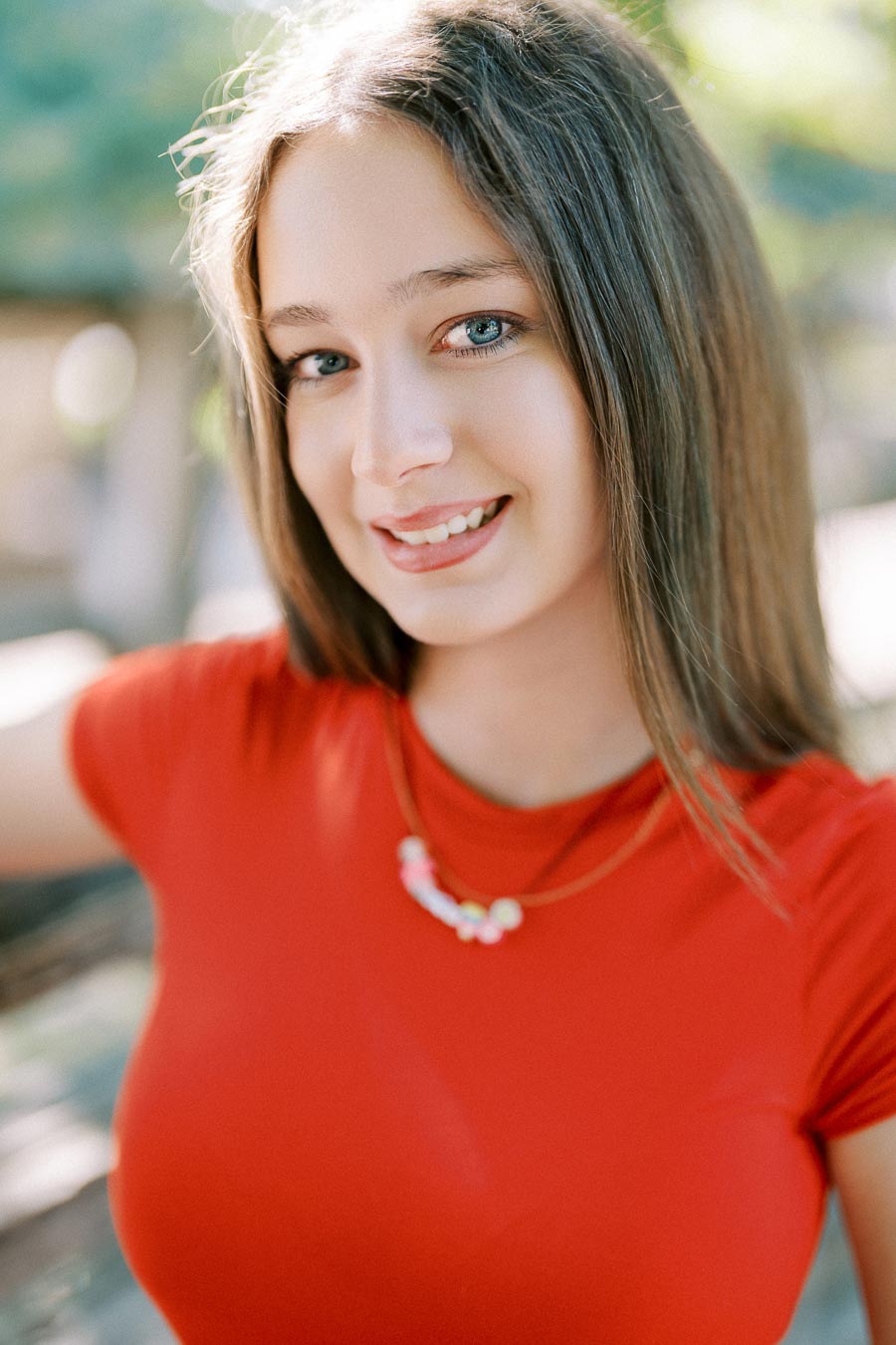 A young woman with long brown hair smiling at the camera, wearing a red shirt and a necklace, in a sunlit outdoor setting.
