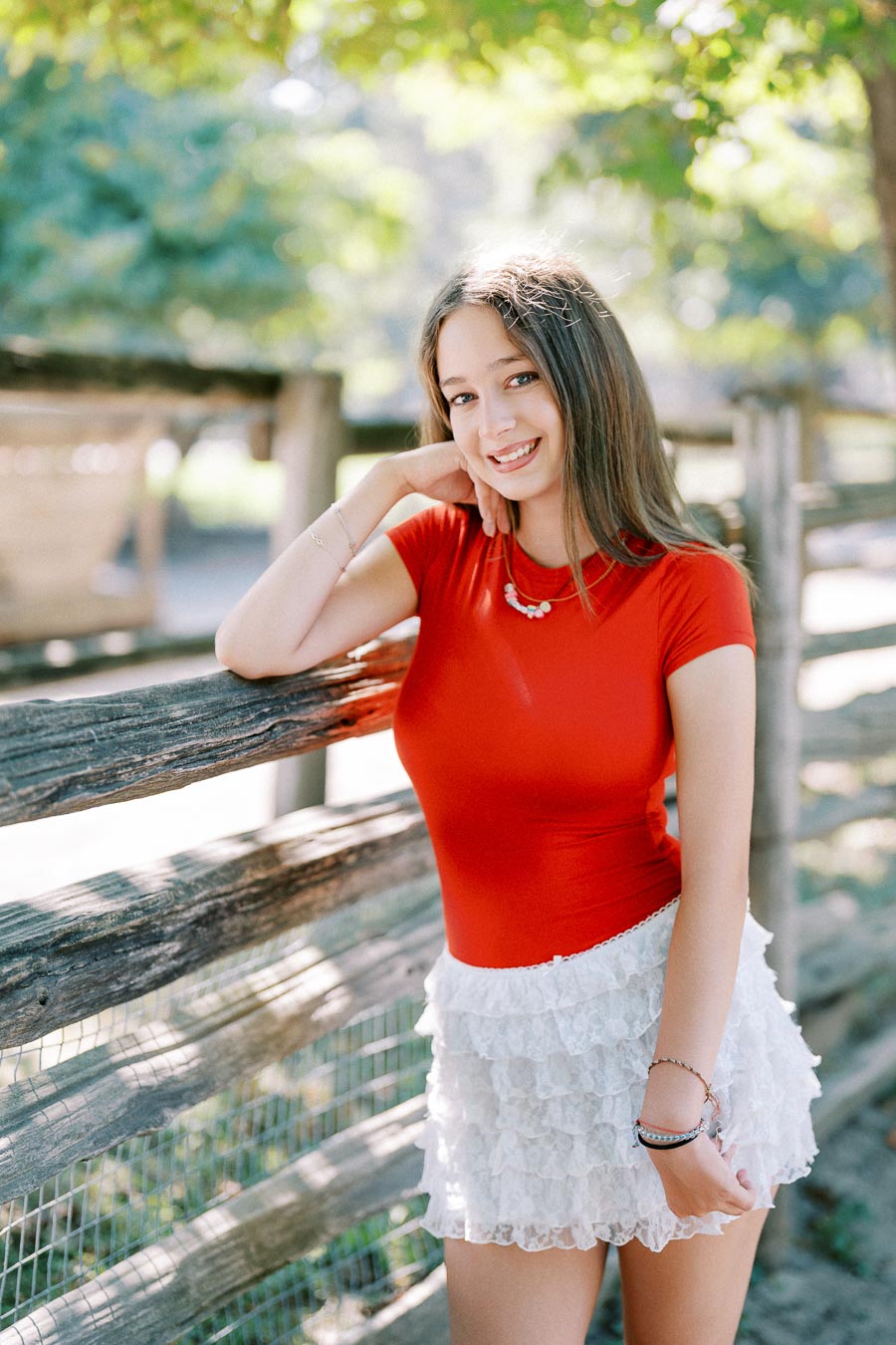 A young woman in a red top and white lace skirt smiling while leaning on a rustic wooden fence, with trees in the background on a sunny day.