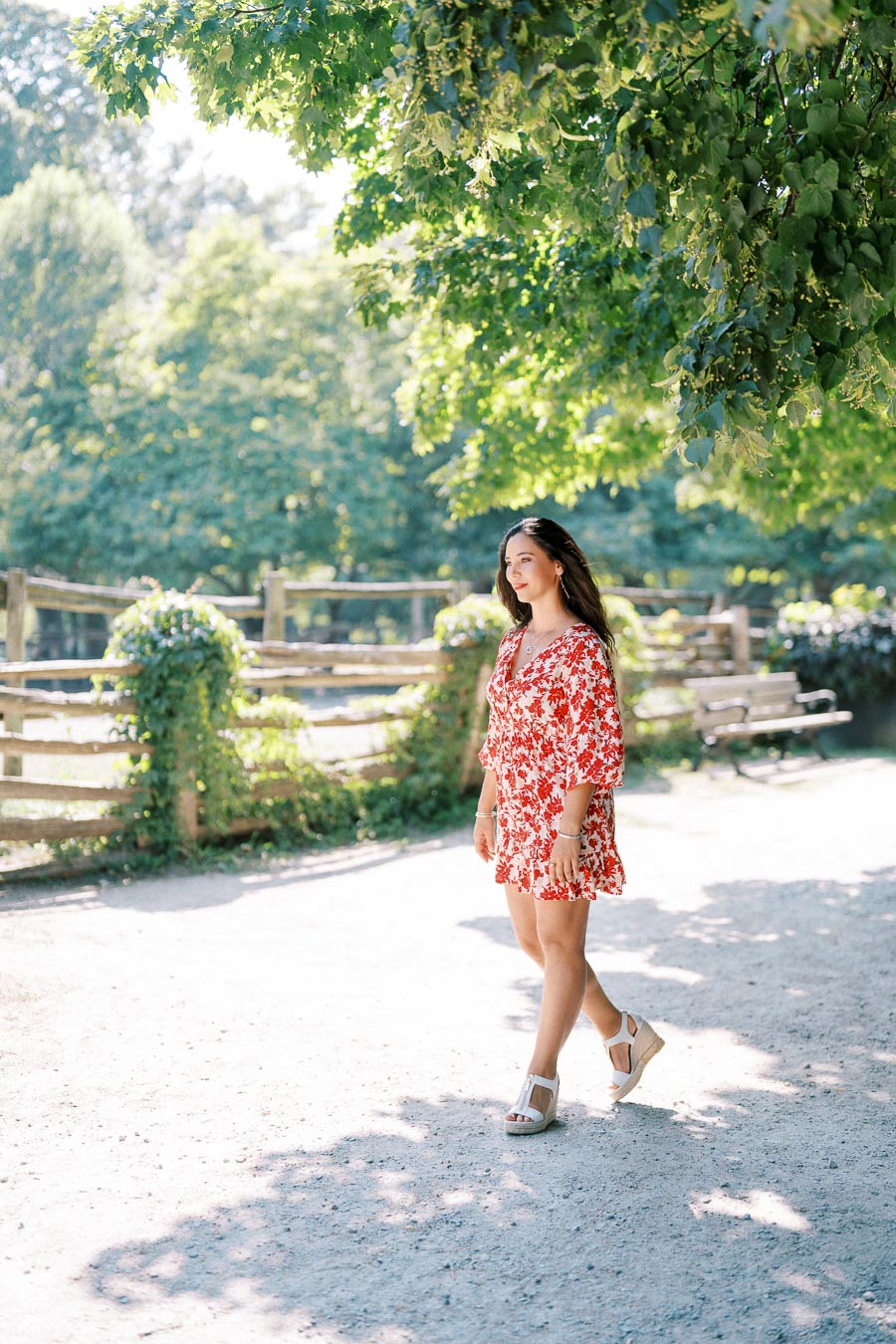 Young woman in a red floral dress walking on a sunny path with trees and a wooden fence in the background.