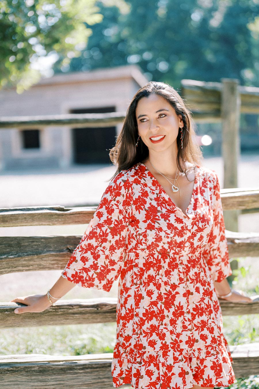 A woman in a red and white floral dress stands by a wooden fence, smiling, with a blurred background of trees and a barn.