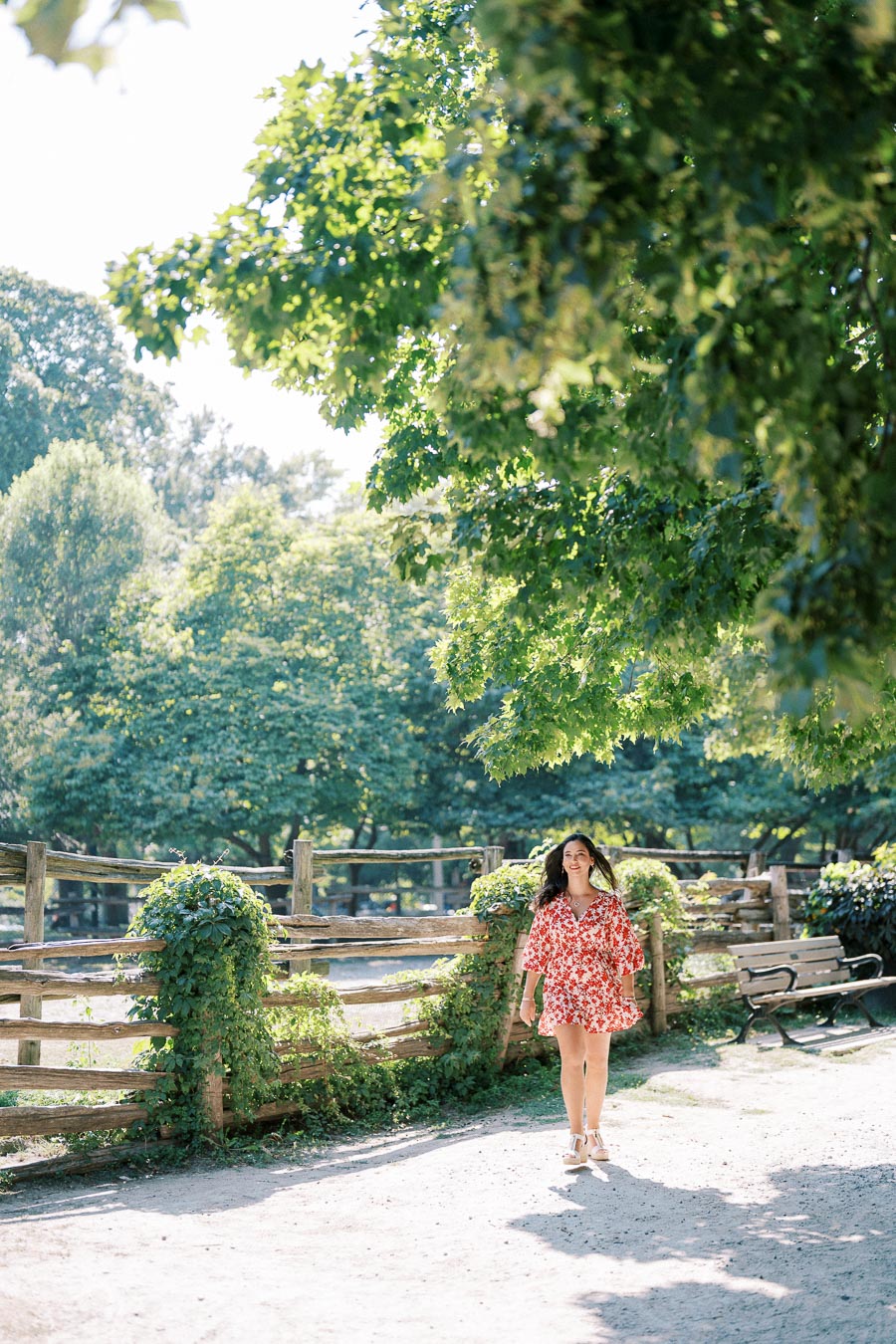 A woman in a floral red dress walks along a sunlit path in a park, surrounded by lush green trees and wooden fences.