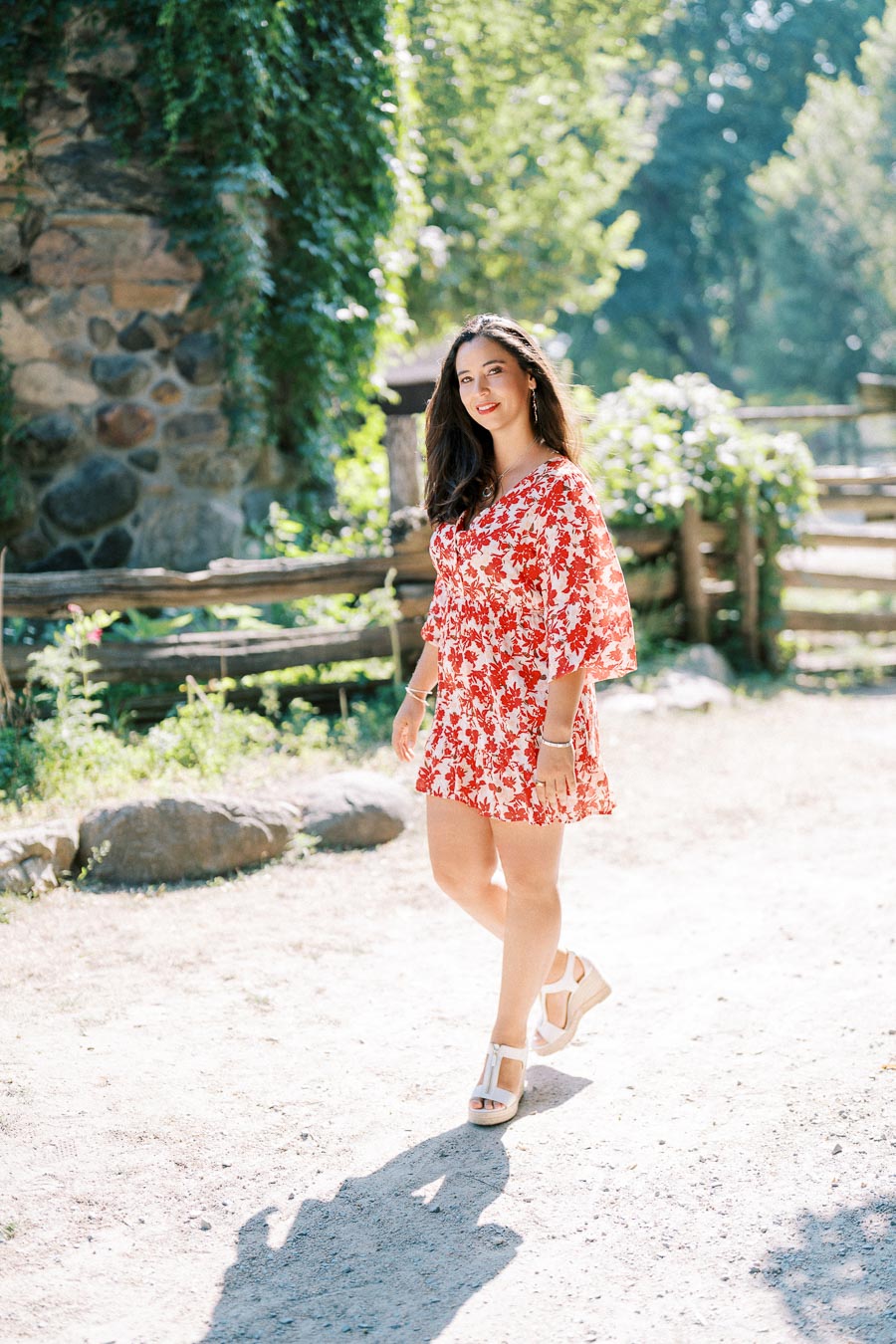 Fashionable woman in a red floral dress walking outdoors on a sunlit path, with a rustic stone wall and greenery in the background.