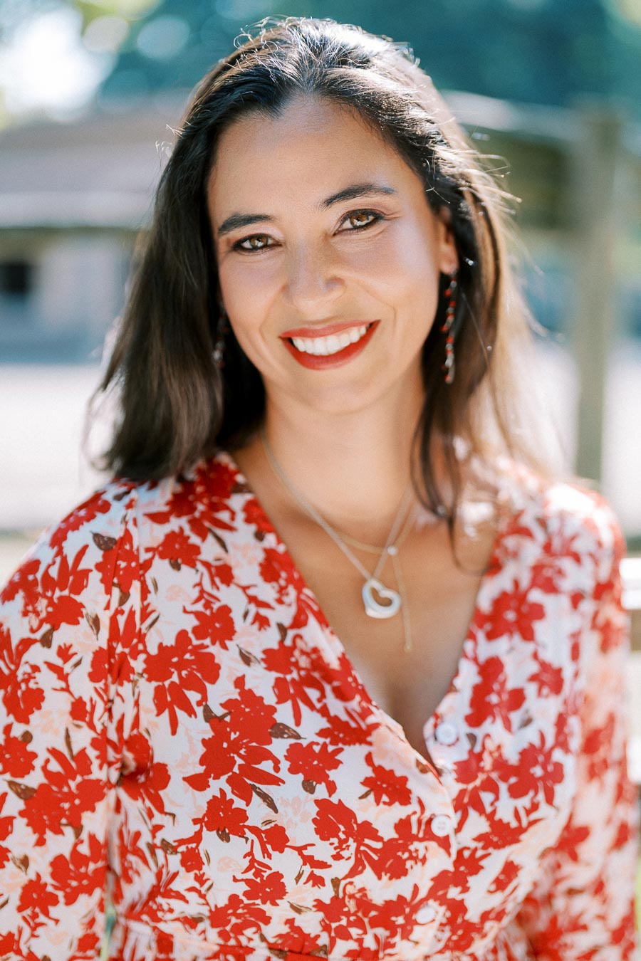 ```markdown Alt text: Woman in a red floral dress smiling outdoors, wearing a heart-shaped necklace and earrings, with blurred greenery in the background. ```
