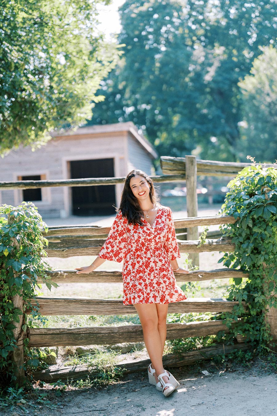 A woman in a red and white floral dress leans against a rustic wooden fence, surrounded by lush greenery, with a wooden barn in the background on a sunny day.