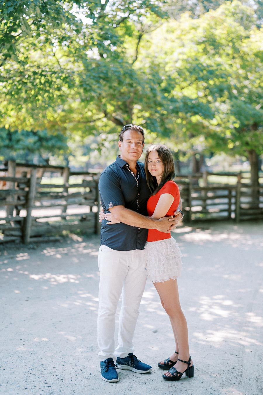A couple embraces in a sunlit park, surrounded by lush green trees and a rustic wooden fence, capturing a moment of love and tranquility.