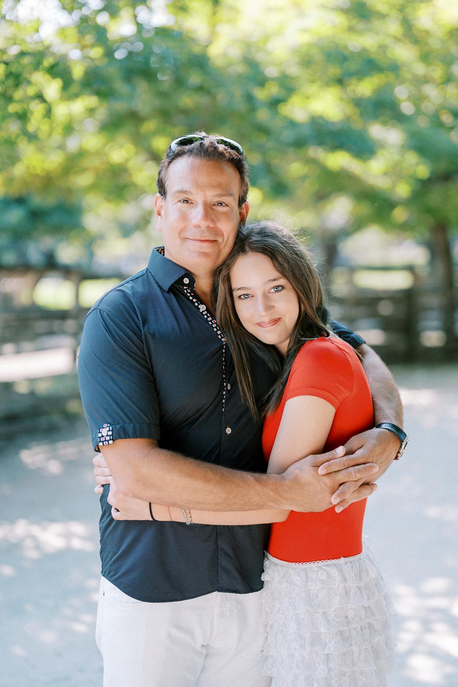 Father and daughter embracing in a sunny park, with green trees in the background, wearing casual summer clothes.